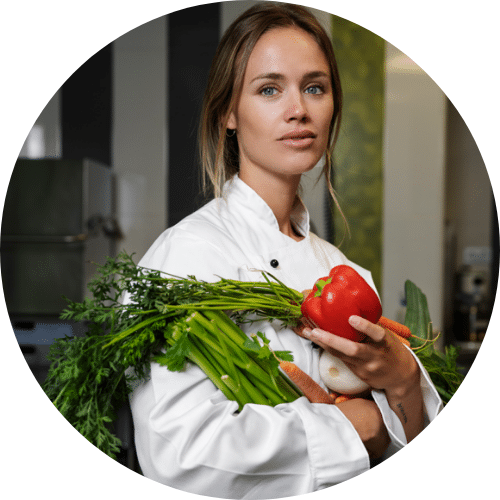 About me 1 Female chef in a white coat and black apron holding fresh vegetables, including carrots, celery, red bell pepper, and onions, standing in a professional kitchen.